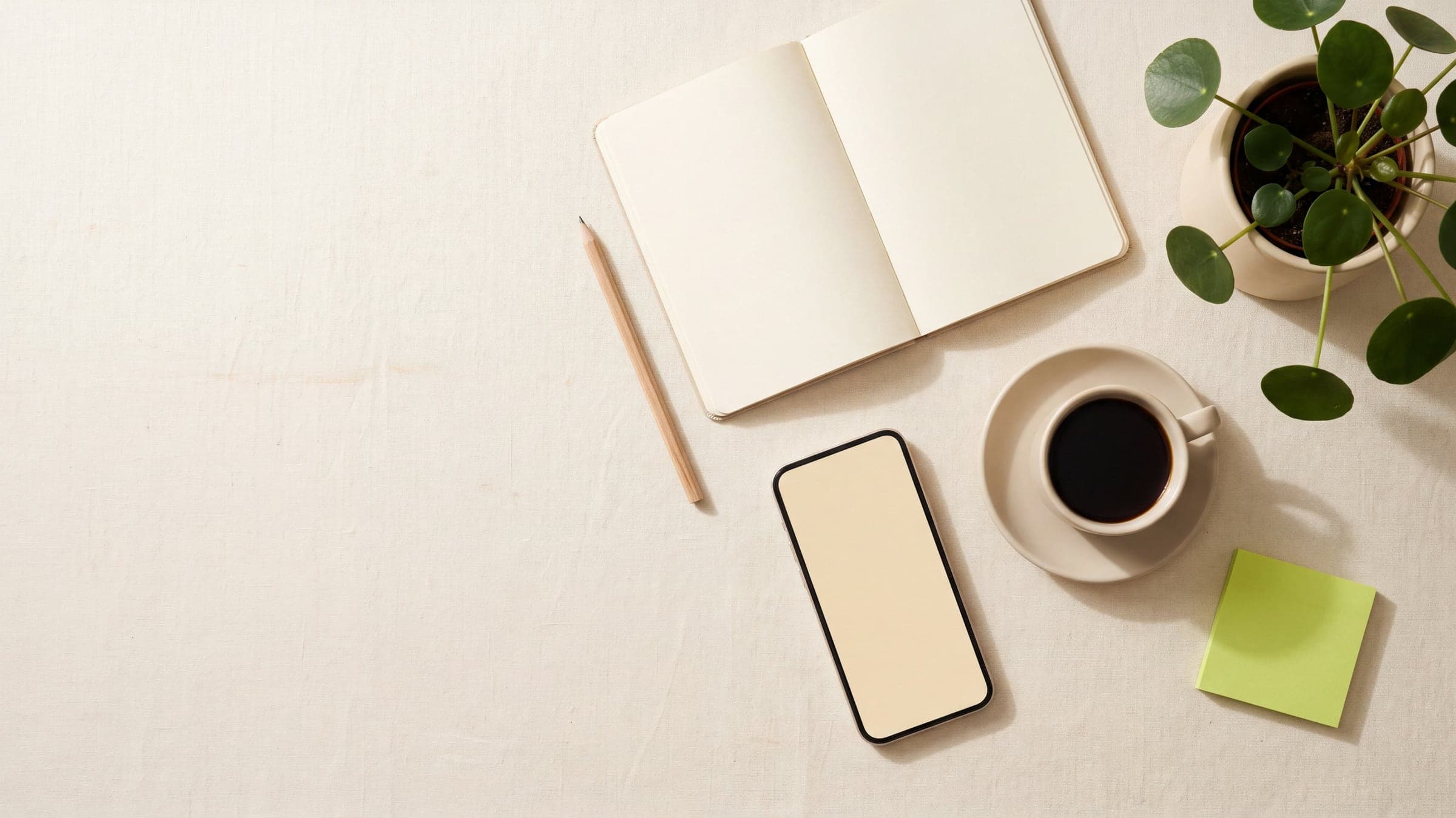 A calm desk from above: an open notebook, a pencil, a phone, a cup of coffee, a small plant and a blank lime sticky note.